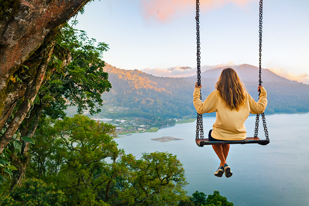 Summer vacation. Young woman sit on tree rope swing on high cliff above tropical lake. Happy girl looking at amazing jungle view. Buyan lake is popular travel destinations in Bali island, Indonesia