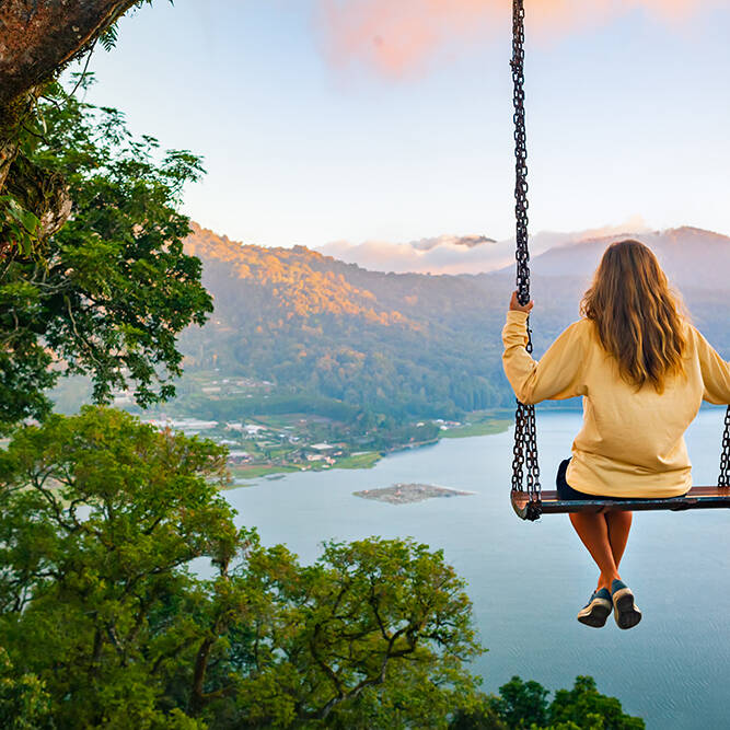 Summer vacation. Young woman sit on tree rope swing on high cliff above tropical lake. Happy girl looking at amazing jungle view. Buyan lake is popular travel destinations in Bali island, Indonesia